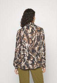 Frau mit lockigem Haar, die eine langärmlige Bluse mit abstrakten braunen, schwarzen und weißen Mustern trägt, kombiniert mit olivgrünen Hosen, und von hinten blickt.
