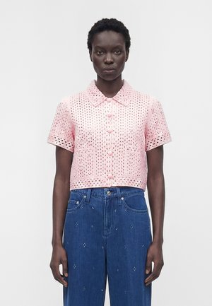 Femme portant un chemisier rose à manches courtes en tissu œillet, boutonné, associé à un jean bleu taille haute avec de petites décorations.