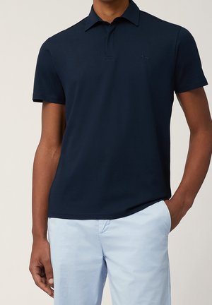 Homme portant un polo bleu marine foncé ajusté et un pantalon bleu clair, une main dans la poche, sur un fond clair uni.