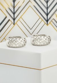 Två metalliska silverringar som visas på en vit pelare. Ringarna har en öppen gallerdesign med geometriska mönster.