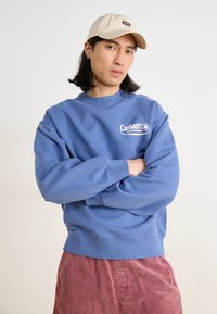 Sweatshirt azul de algodão com punhos e bainha canelados, apresentando um logótipo branco. Usado com calções de veludo-canelado vermelhos e um boné bege.