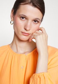 Blusa laranja, texturizada, com decote redondo e detalhes franzidos. A modelo usa brincos de argola dourados e vários anéis nos dedos.