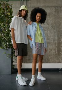 Kaksi mallia seisoo vierekkäin. Mies pukeutuu valkoiseen ylisuurikokoiseen paitaan ja ruskeisiin shortseihin; nainen pukeutuu siniseen ylisuurikokoiseen paitaan ja raidallisiin shortseihin. Molemmilla on valkoiset tennarit jalassa.