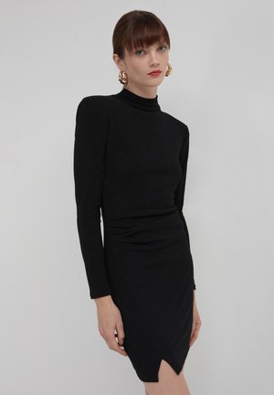 Frau mit glatten braunen Haaren, die goldene Hörnerohrringe und ein enges schwarzes Langarmkleid mit einem kleinen Schlitz am Saum trägt.