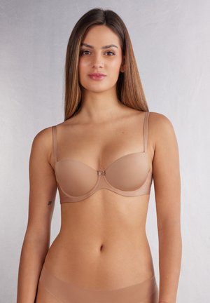 Light beige bra made of smooth, stretchy fabric. Molded cups, thin adjustable straps, and a decorative bow at the center front.