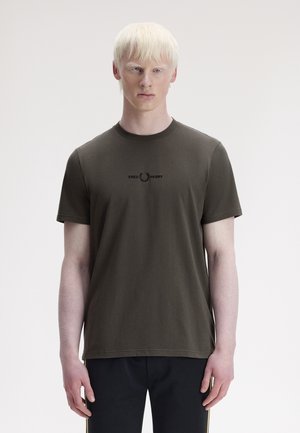Ung mand med bleg hud og blondt hår iført en mørkegrøn Fred Perry t-shirt og sorte bukser foran en hvid baggrund.