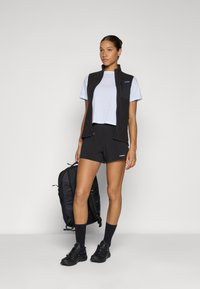 Zwarte ritssluiting vest over een lichtgrijs T-shirt, gecombineerd met zwarte shorts. Draagt zwarte schoenen en marineblauwe sokken, en houdt een zwarte rugzak vast.