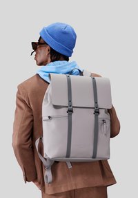 Zaino grigio con patta e spalline grigie, dotato di una tasca frontale con zip. Materiale liscio con un design minimalista.