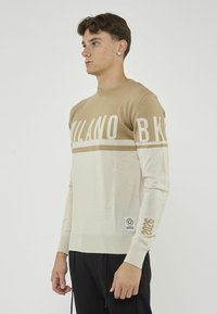 Maglione a maglia beige e crema con la scritta "MILANO BK" stampata sul petto, polsini e orlo a costine; presenta una toppa con logo e un dettaglio "2026".