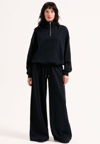 Schwarzes Set aus passendem Sweatshirt und weitem Hosen. Das Sweatshirt hat einen hohen Kragen und eine Fronttasche. Weicher Stoff mit subtiler Textur.
