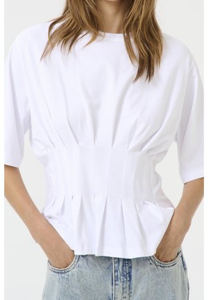 Femme portant un haut blanc à manches courtes avec taille froncée, mains dans les poches d'un jean denim bleu clair, visible du cou aux hanches.