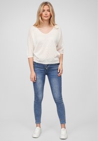 Hvit strikket genser med V-hals og korte ermer, preget av et chevronmønster, kombinert med blå skinny jeans og hvite joggesko.