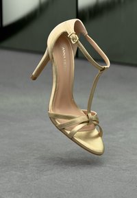 Goldene High-Heel-Sandalette mit T-Riemen und Schnalle, schmalem Absatz und über den Zehen gekreuzten Riemen, präsentiert auf einer grauen Fläche.