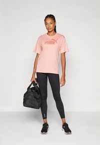 Šviesiai rausva marškinėlių su „PUMA“ logotipu, derinama su juodomis leginsėmis ir juodais sportbačiais. Modelis laiko juodą sporto krepšį su tinklinėmis detalėmis.