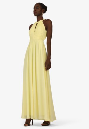 Frau trägt ein ärmelloses, blassgelbes Neckholder-Kleid mit plissiertem Oberteil und langem, fließendem Rock, kombiniert mit schwarzen Sandaletten mit hohen Absätzen.
