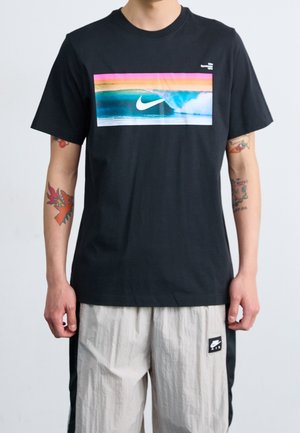Osoba nosząca czarną koszulkę Nike z kolorową falistą grafiką i logo, jasnoszare spodenki Nike z czarnymi panelami bocznymi i naszywką z logo.