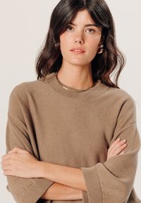 Bruine gebreide trui met een losse fit, brede mouwen en geribbelde halslijn. Het model draagt een eenvoudige gouden ketting en parel oorbellen.