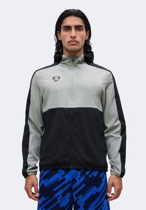 Mann trägt eine grau-schwarze Jacke mit Reißverschluss und Kapuze sowie blaue, gemusterte Sportshorts und steht vor einem einfarbig hellen Hintergrund.