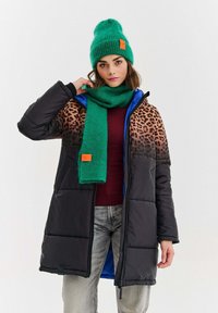 Žena oblečená v čiernej puffrovacej bunde s tričkom s leopardím vzorom, zelenou pletenou čiapkou a zodpovedajúcim šálom, bordovom topu a vyblednutých šedých džínsoch.