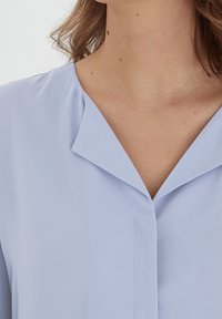 Blouse bleu clair avec un col, texture douce, fermeture à boutons sur le devant et coupe décontractée. Présente un design délicat en v.