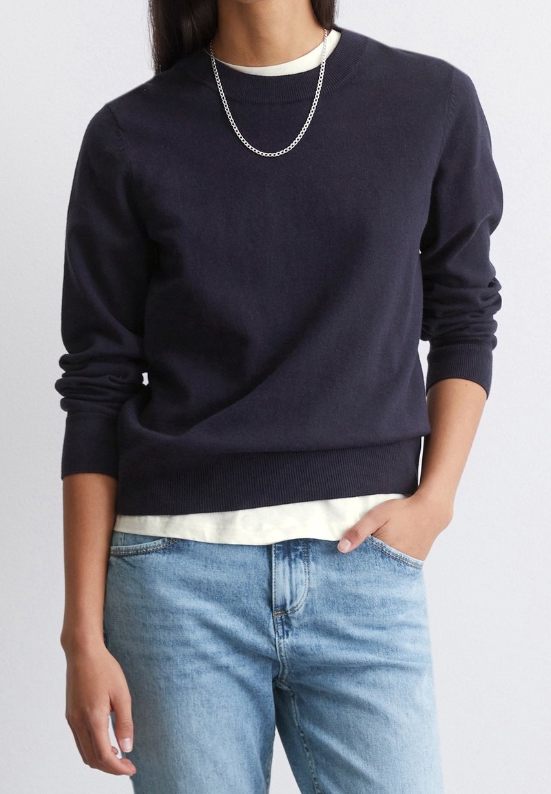Personne portant un pull bleu marine foncé sur une chemise blanche, un jean bleu clair, un collier chaîne argenté, une main dans la poche et l'autre détendue le long du corps.