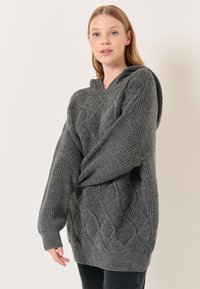 Maglione grigio lavorato a maglia con motivo a treccia, vestibilità over-size e cappuccio. La texture è a coste, con un aspetto morbido e spesso del filato.