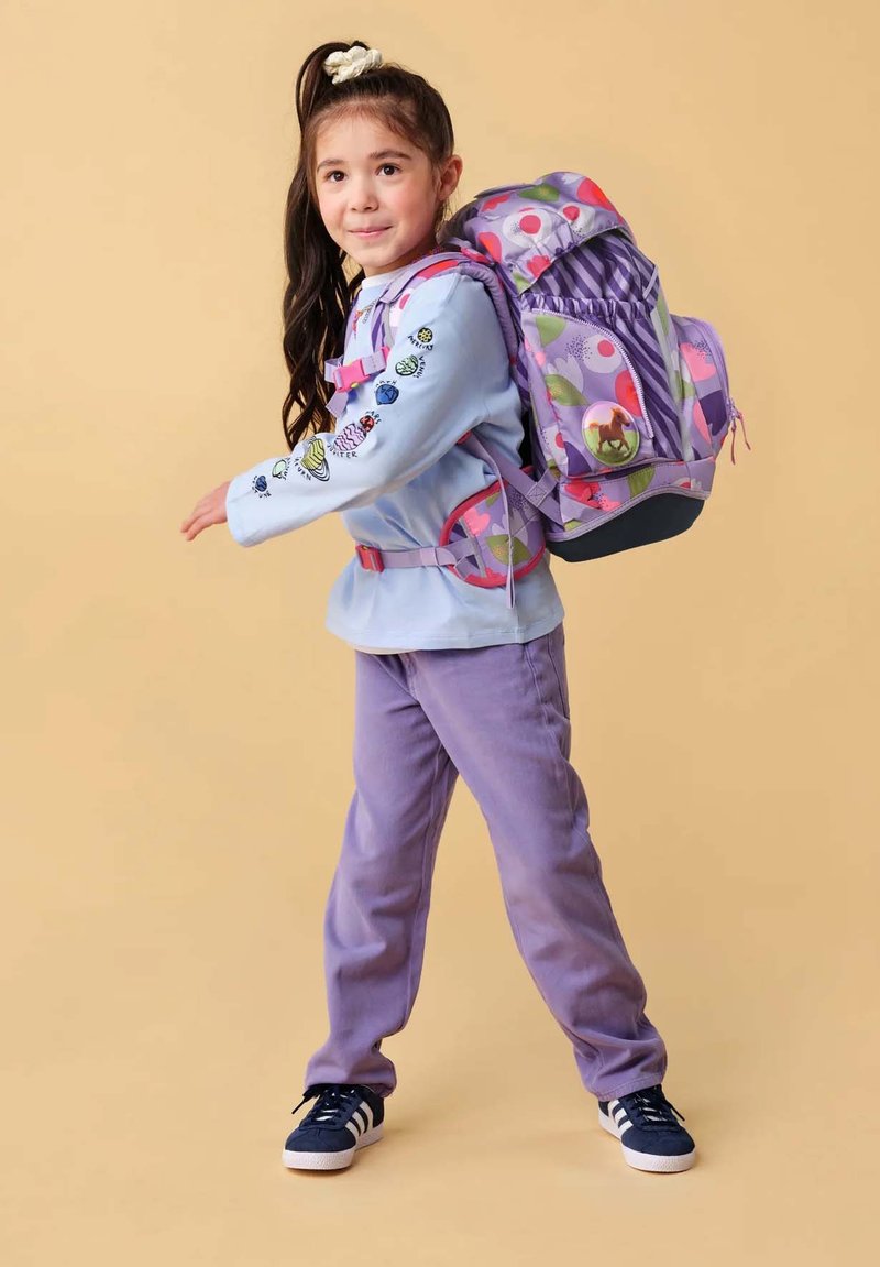 Junges Mädchen, das einen bunten Rucksack trägt, mit violetten Hosen und einem hellblauen Langarmshirt, geht vor einem beigen Hintergrund.