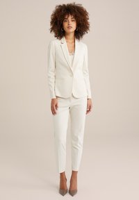 Costume blanc sur mesure composé d'un blazer ajusté et d'un pantalon slim, fabriqué à partir d'un tissu lisse. Le blazer est doté d'un bouton unique et de revers crantés.