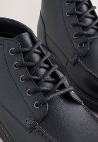 Schwarze Leder-Stiefeletten mit strukturierter Oberfläche, ausgestattet mit runden Schnürsenkeln, Metallösen und kontrastierenden Nähten entlang der Zwischensohle.