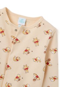 Un body de bebé en color beige claro con un patrón repetido de ositos de dibujos animados con camisetas rojas, fabricado con material suave, cuello redondo y cierre a presión.