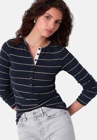 Chemise henley à manches longues bleu marine avec des rayures horizontales blanches, dotée d'une patte de boutonnage et d'une texture côtelée, associée à un jean gris clair.