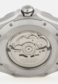 Montre en acier inoxydable avec un mouvement mécanique visible comprenant des engrenages et des pierres précieuses. Comprend des détails de marque et une couronne hexagonale.