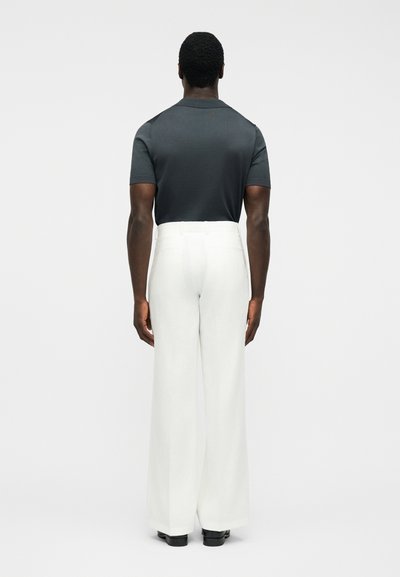 Muž v krátkém rukávu tmavě šedé polokošili zastrčené do vysokých širokých bílých kalhot, stojící zády.