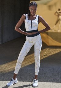 Weißes Sportset mit goldenen Marmor-Mustern; ärmelloses Top mit Frontreißverschluss, hoher Bund und Leggings; kombiniert mit schwarz-weißen Turnschuhen.