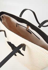 Sac cabas tissé crème avec poignées en cuir noir, applique en forme de chauve-souris noire, intérieur en tissu beige et poche zippée avec fermoir doré.