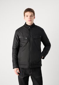 Schwarze Zip-Up-Jacke mit hohem Kragen, zwei Brusttaschen mit Druckknöpfen und einer glatten, leicht glänzenden Textur. Getragen mit dunklen Jeans.