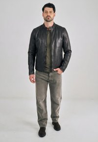 Mężczyzna stojący w czarnej skórzanej kurtce, oliwkowozielonej koszulce, szarych dżinsach i ciemnobrązowych butach na jasnym, jednolitym tle.