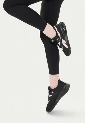Nogi w czarnych leginsach, różowych skarpetkach i czarnych butach sportowych Reebok z różowymi akcentami, jedna noga zgięta i uniesiona na jednolitym jasnym tle.