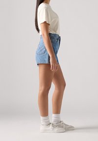 Högmidjade blå denimshorts med rak skärning, kombinerat med en krämfärgad t-shirt och vita sneakers med vita strumpor. Slät textur.