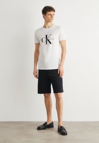 Balta kokvilnas t-krekls ar melno "CK" logotipu un tekstu "Calvin Klein Jeans", kombinēts ar melnajiem šortiem un melnajiem mokasīniem uz neitrāla fona.