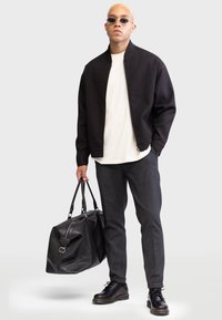 Veste bomber noire texturée sur un t-shirt blanc, pantalon gris et chaussures en cuir noires. Tenant un grand sac de voyage noir.