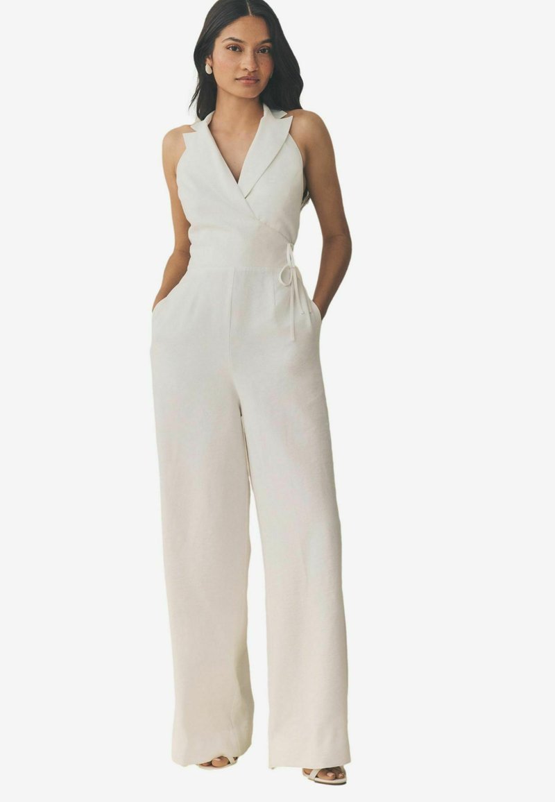 Jumpsuit blanc sans manches avec une taille cintrée, un design à jambes larges, un col à revers, des poches latérales et un détail en tissu à la taille.