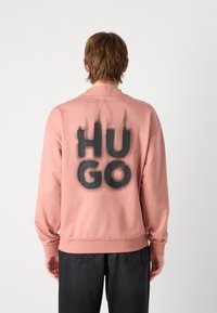 Puhaszövetből készült, hosszú ujjú, kerek nyakú pulóver, lágy rózsaszín árnyalatban, a hátsó részen nagy, fekete "HUGO" felirattal, füstös átmeneti hatással. Texturált felület.