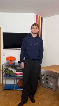 Νέος άνδρας με μπλε φούτερ με κουκούλα και μαύρο παντελόνι στέκεται σε σαλόνι με τηλεόραση, πορτοκαλί λάμπα, φυτό, βιβλία, καλαθοσφαίριση και βαλίτσα με αυτοκόλλητα.