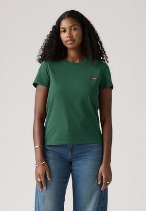 Femme aux cheveux bouclés portant un t-shirt vert et un jean bleu, debout devant un fond clair uni.