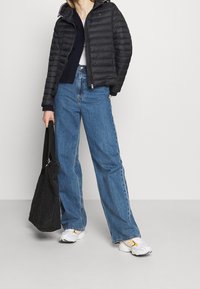 Giacca a vento nera con cappuccio, jeans denim a gamba larga azzurri, sneakers bianche con accenti arancioni e una borsa a tracolla nera trapuntata.