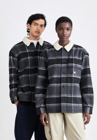 Schwarz- und graue karierten Jacken mit einem weichen weißen Kragen, ausgestattet mit Knöpfen und einer Brusttasche mit einem Logo. Getragen von zwei Modellen.