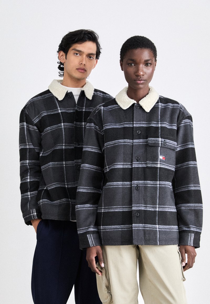 Schwarz- und graue karierten Jacken mit einem weichen weißen Kragen, ausgestattet mit Knöpfen und einer Brusttasche mit einem Logo. Getragen von zwei Modellen.