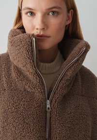 Braune Fleecejacke mit hohem Kragen, ausgestattet mit einem durchgehenden Reißverschluss und strukturierter Oberfläche, getragen über einem beigen Strickpullover.