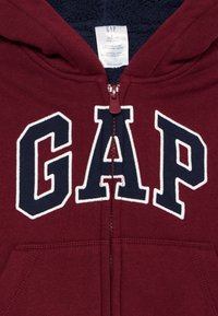 Felpa con zip color mogano con logo "GAP" in lettere bianche e blu navy; collo foderato in pile; tasche frontali; texture del tessuto morbida.